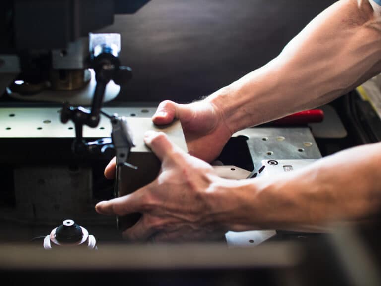 Close-up of hands operating a metalworking machine, with tools and metal components visible in a workshop setting.